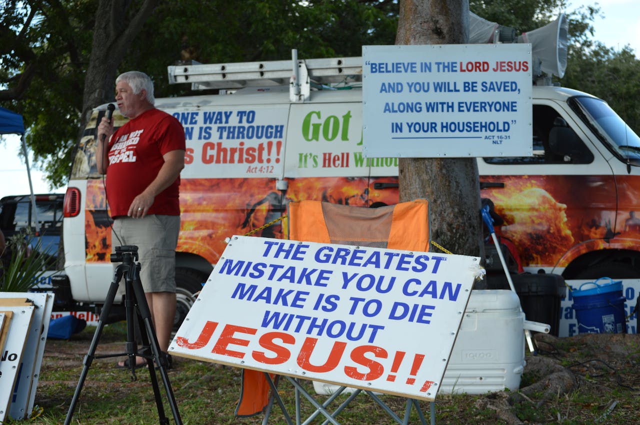 A man engages in outdoor evangelism with religious signs and a microphone by a van.