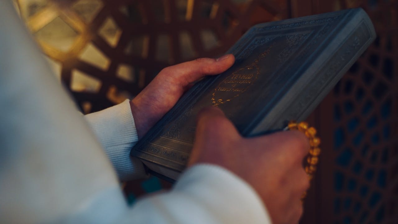 Close-up of hands holding a Quran and prayer beads inside a mosque.