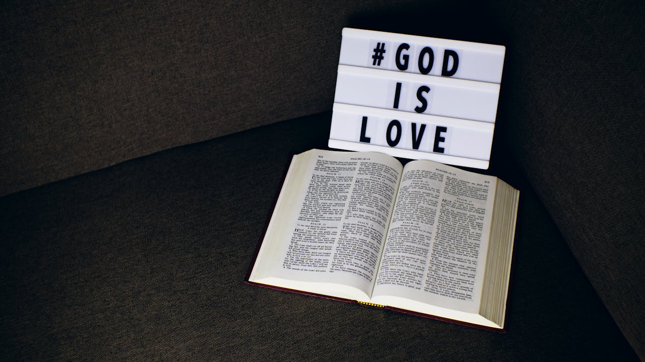 Open Bible placed beside a lightbox sign reading 'God is Love' on a dark fabric surface.
